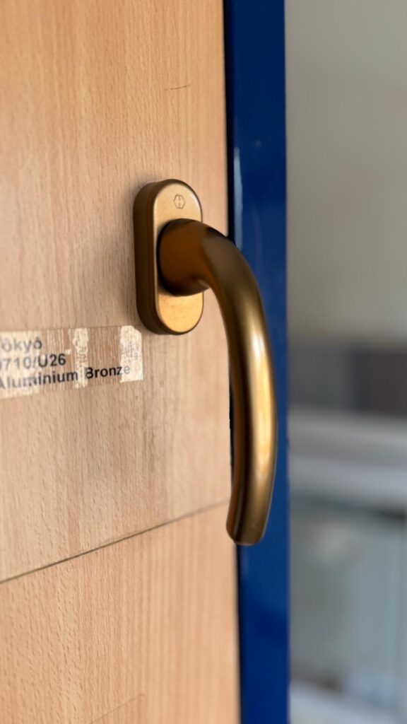 A lever handle in aluminium bronze on a woodgrain showroom sample board, beside its label